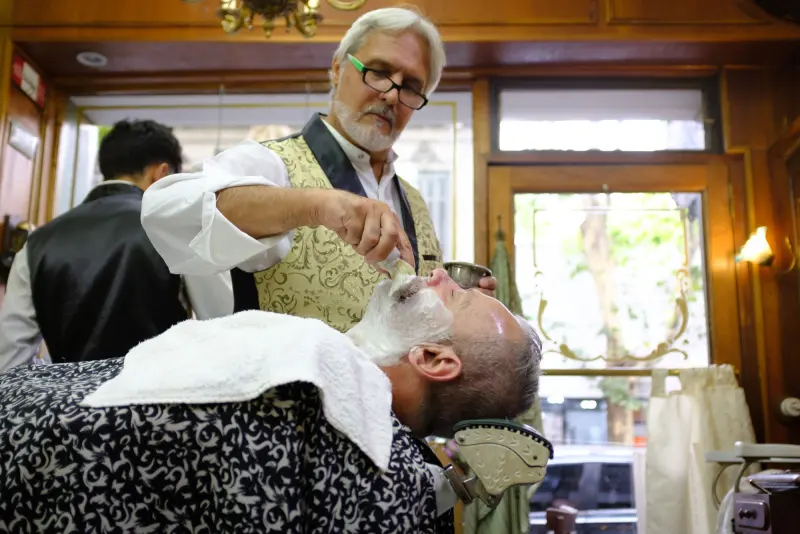 Julio Rivas afeitando con navaja a un cliente con toalla en el pecho